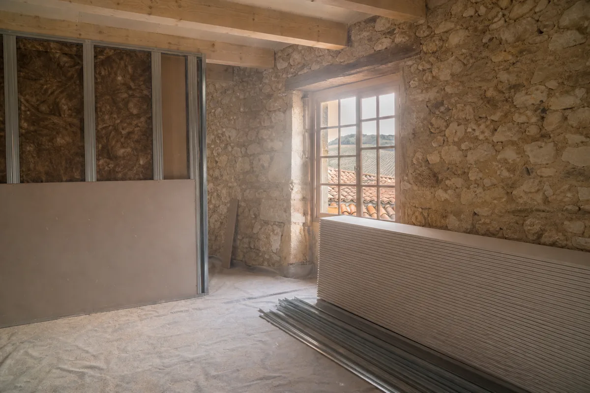 Plaques de plâtre et rails métalliques appuyés contre un mur en moellons calcaires dans une maison ancienne du Périgord