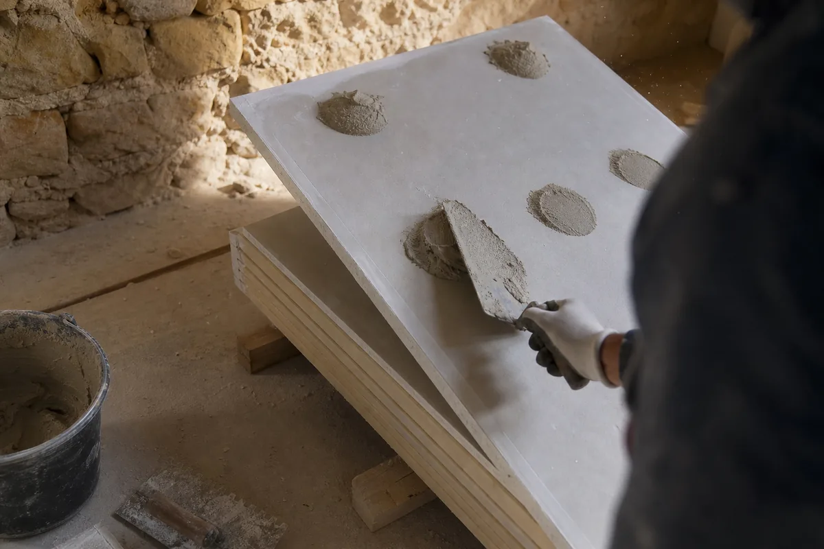 Plaques de plâtre contrecollées sur isolant, stockées contre un mur en moellons calcaires avant pose